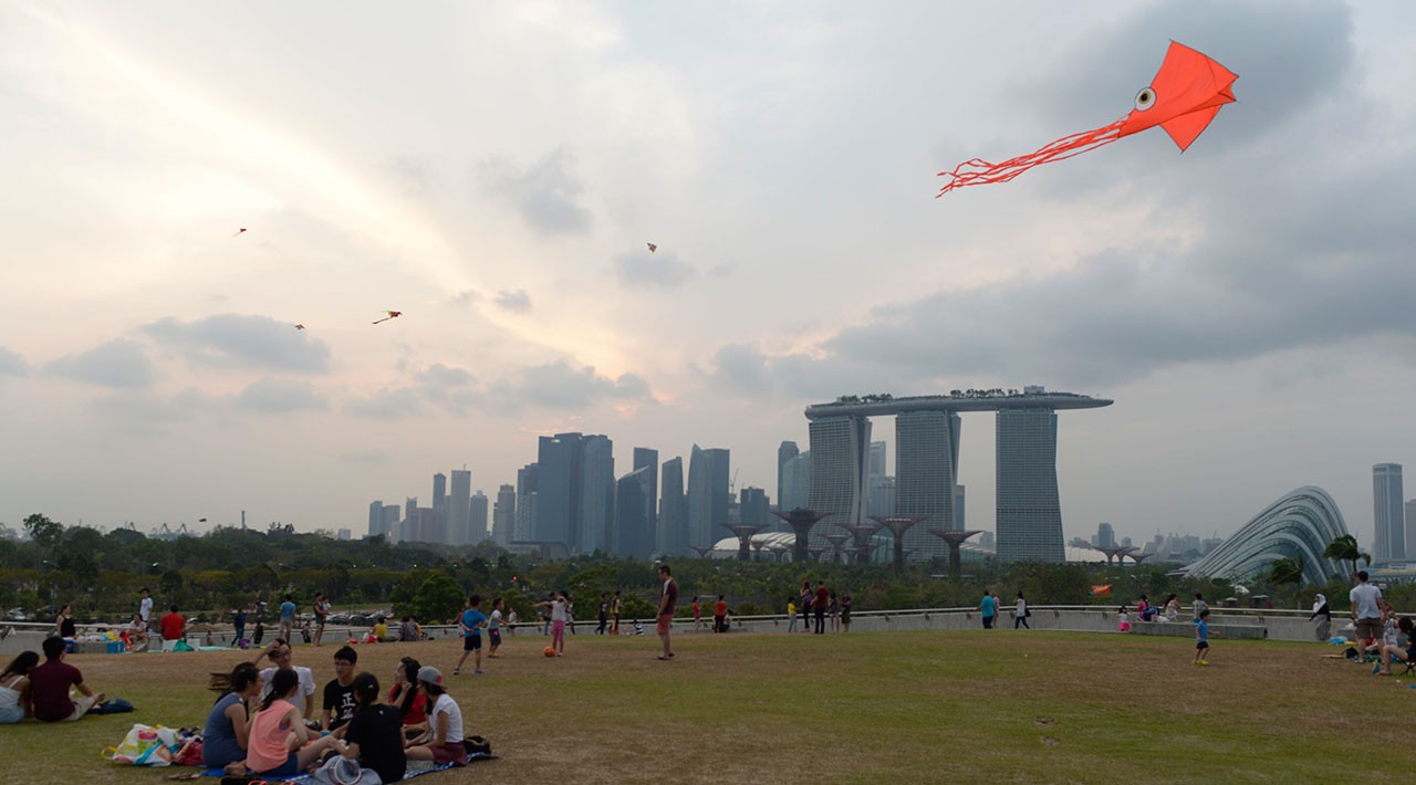An orange kite flies over a sunny park where people enjoy the skyline of Singapore in the distance. 